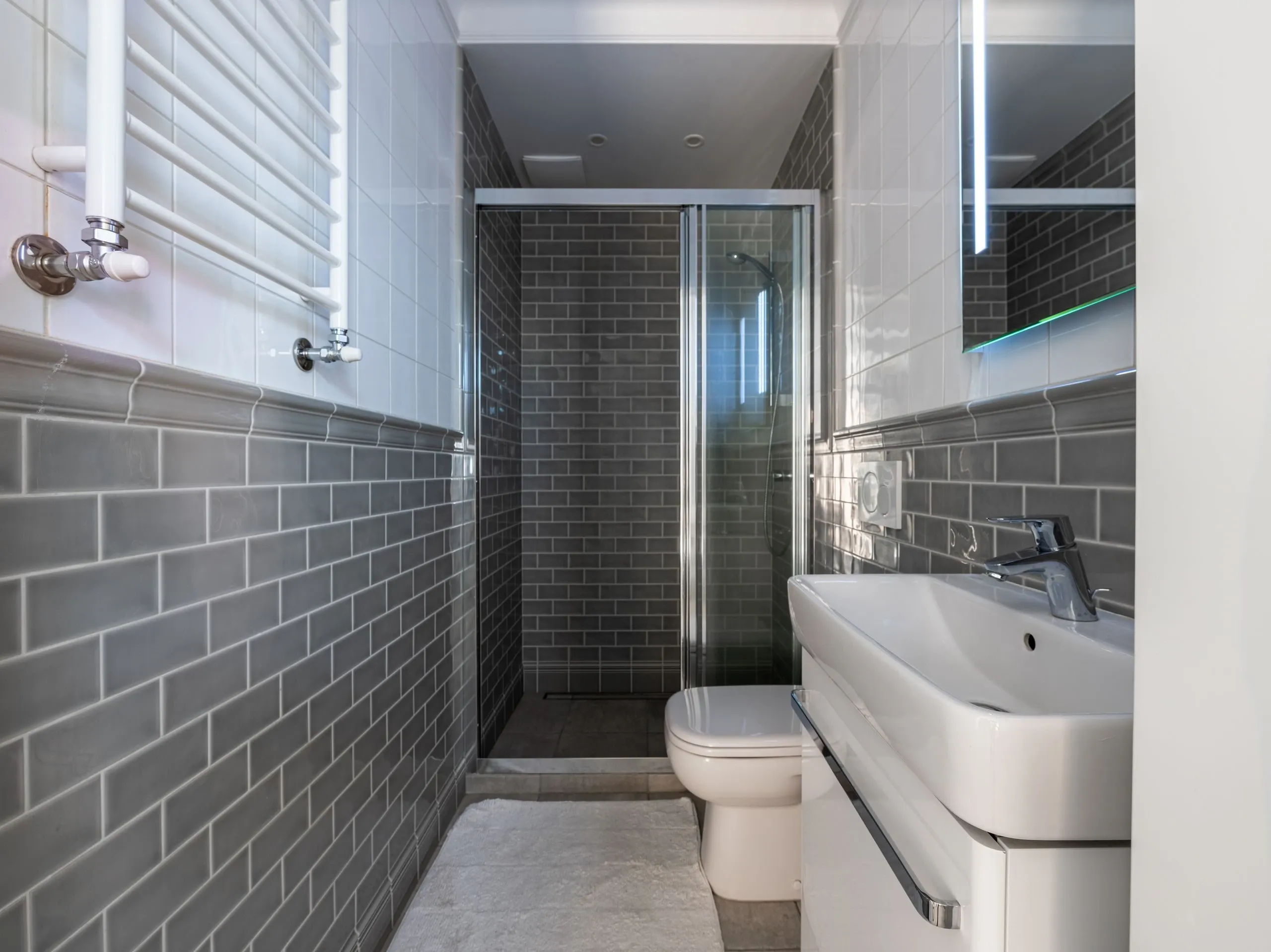 Une salle d'eau étroite en carrelage gris : douche au fond, vasque et sèche-serviettes